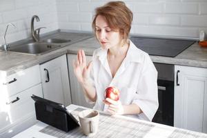 contento mujer utilizando digital tableta para vídeo llamada amigos y padres. mujer con taza de café utilizando ordenador portátil en cocina foto