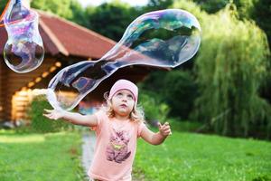 contento niña jugando con jabón burbujas exterior. un pequeño niña estallidos un jabón burbuja foto