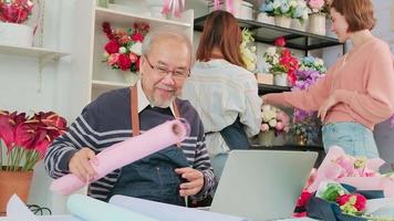 el anciano asiático propietario de una floristería vive en línea transmitiendo para vender en un sitio web de comercio electrónico, empleado y cliente con flores detrás, trabajando en una floristería colorida, un pequeño empresario. video