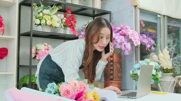 een jong Aziatisch vrouw bloemist baasje, werken met laptop, verkoop bloemen afspreken, pratend Aan mobiel telefoon in kleurrijk bloem winkel op te slaan met een mooi bundel van bloesems, en e-commerce bedrijf. video