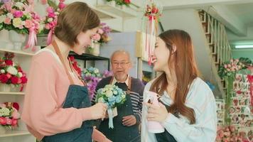 Young female florist workers have fun teasing together, and a senior Asian male owner shows a beautiful bunch of blossoms, happy work in a colorful flower shop store, SME business entrepreneur people. video