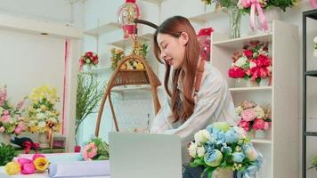 un Jeune asiatique femelle fleuriste démontre et spectacles floral décorer via en ligne vivre diffusion avec portable dans coloré fleur magasin boutique avec magnifique grappes de fleurir, et commerce électronique entreprise.