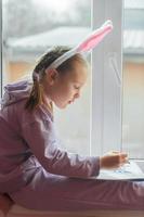 a schoolgirl girl with rabbit ears sits on the window and draws a rabbit in her album, preparing for Easter photo