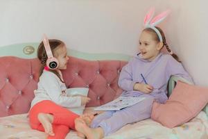 two sister girls have fun at home, draw a bunny and listen to music in headphones, the older girl has a hairband with rabbit ears on her head, waiting for the Easter holiday photo