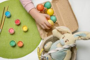 child puts painted Easter eggs in a basket, next to a brush and paint and an Easter bunny photo