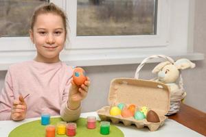 happy pretty girl sitting at a table with paints and an easter bunny shows a painted easter egg photo