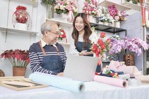 Asian senior male florist owner discusses with young beautiful female employee about bunch of fresh blossom arrangements for online business, happy work in colorful flower shop store, e-commerce SME. photo