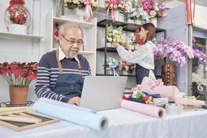 el anciano propietario asiático de una floristería que trabaja con una laptop en un sitio web para negocios en línea, una joven y hermosa comerciante que organiza flores frescas detrás, una colorida tienda de flores, una pyme de comercio electrónico. foto