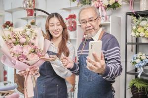 E-commerce business, senior Asian male florist worker and daughter show floral arrangements, online live streaming, selfie by smartphone application in bright flower shop, beautiful blossoms store. photo