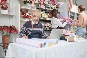 Asian senior male florist owner works with laptop, online selling in e-commerce business website, beautiful female employee and customer with blossom, colorful flower shop store, SME entrepreneur. photo