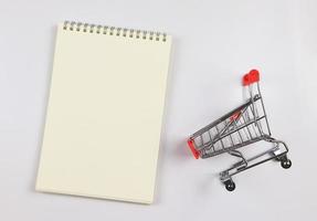 flat lay of  blank page opened notebook and shopping trolley or shopping cart  on white background. shopping list concept. photo