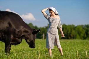A young girl in a cow pasture is standing in a dress and a kerchief , looking at a cow photo