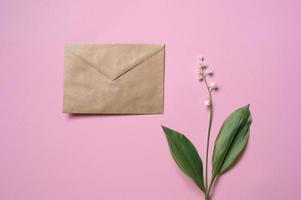 Sprigs of lily of the valley flowers lie on a pink background next to the envelope photo