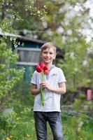 A cute boy in a white T-shirt stands with tulips near a green tree photo