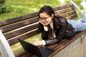 A young girl with glasses is lying on a bench with a laptop and working photo