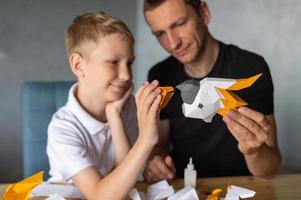 un linda chico es sentado con su papá a el mesa y coleccionar origami. pegamento el partes foto