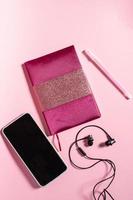 A pink notebook with a pen and headphones lies on a monochrome background next to the phone photo