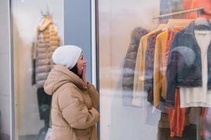 un mujer en un blanco sombrero y Saco cubre su boca con su mano y mira a el tienda ventana foto