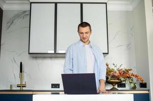 A young man in a shirt works from home on a laptop while standing in the kitchen photo