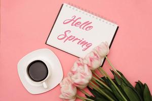 Text HELLO SPRING next to a beautiful bouquet of delicate pink tulips and a cup of coffee on a pink background. There may be your text. photo