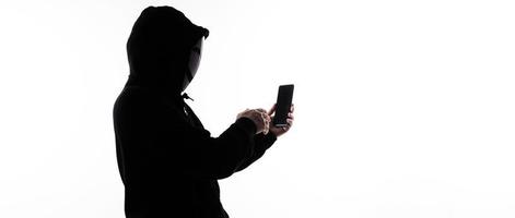 Hacker Anonymous and face mask with smartphone in hand. Man in black hood shirt holding and using mobile phone on white background. Represent cyber crime data hacking or stealing personal data concept photo