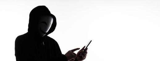 Hacker Anonymous and face mask with smartphone in hand. Man in black hood shirt holding and using mobile phone on white background. Represent cyber crime data hacking or stealing personal data concept photo