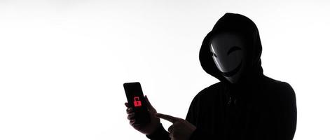 hacker anónimo y mascarilla con smartphone en la mano. hombre con camisa de capucha negra sosteniendo y usando un teléfono móvil con fondo blanco. representan el concepto de piratería informática o robo de datos personales foto