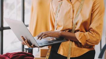 Fashion designer l young asian woman working using laptop, tablet and smiling while standing in workshop Responding on business video