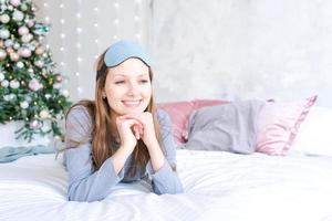 Cheerful girl in sleeping eye mask lying in bed in blue pajamas against photo