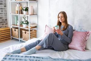 Attractive brunette woman is sitting on bed in her pajamas drinking hot tea or photo