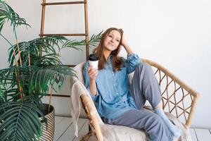 Sweet happy woman sits alone on an armchair in front curtains and looks at view photo