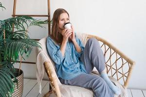 Sweet happy woman sits alone on an armchair in front curtains and looks at view photo