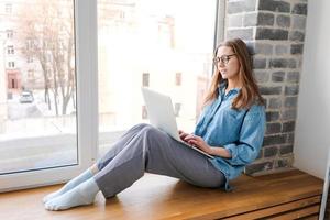Happy casual beautiful woman in glasses working on laptop sitting on windowsill photo