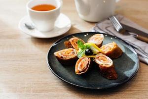 Rolls of thin pancakes with smoked salmon and cream cheese on dark plate on wooden table. Delicious and healthy breakfast or brunch. photo