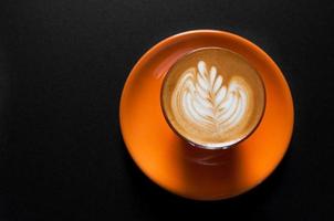 Hot latte in orange cup with floral pattern in foam on a dark background photo