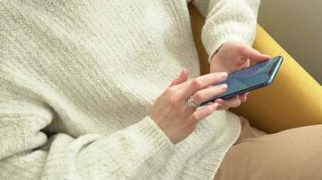 Young woman holding and using mobile phone - typing text on the screen and surfing the internet. Girl sitting relaxed in yellow chair, hold smartphone working, communicate, read and watching video