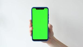 Hand of young woman holding a smartphone with vertical green chroma key screen on gray backdrop. 4k template. Girl hands keeps cell phone with green mockup screen background in front of the camera video