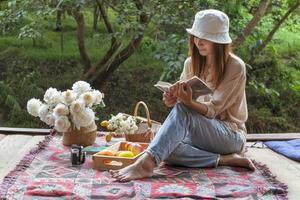 Woman in hat and old-fashioned dress sits and reads a book on a wooden balcony against a green forest backdrop. photo