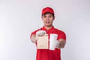 Image of a happy young delivery man in red cap blank t-shirt uniform standing with empty brown craft paper packet isolated on light gray background studio photo