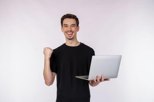 Portrait of young handsome smiling man holding laptop in hands, typing and browsing web pages while doing a winning closed fist gesture isolated on white background photo