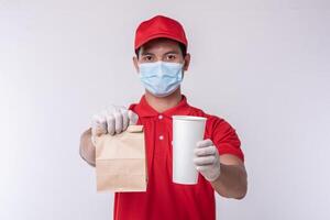 Image of a happy young delivery man in red cap blank t-shirt uniform face mask gloves standing with empty brown craft paper packet isolated on light gray background studio photo