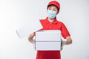 imagen de un joven repartidor consciente con gorra roja en blanco, camiseta uniforme, guantes de máscara facial de pie con una caja de cartón blanca vacía aislada en un estudio de fondo gris claro foto