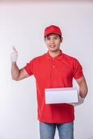 imagen de un joven repartidor feliz con gorra roja en blanco uniforme de camiseta de pie con una caja de cartón blanca vacía aislada en un estudio de fondo gris claro foto