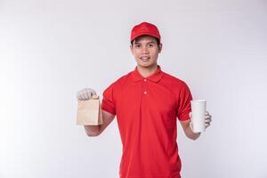 imagen de un joven repartidor feliz con gorra roja en blanco uniforme de camiseta de pie con un paquete de papel artesanal marrón vacío aislado en un estudio de fondo gris claro foto