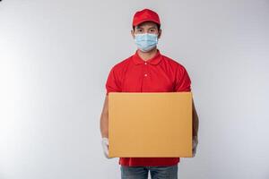 imagen de un joven repartidor consciente con gorra roja en blanco camiseta uniforme guantes de máscara facial de pie con una caja de cartón marrón vacía aislada en un estudio de fondo gris claro foto