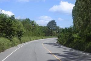 Country asphalt road in Thailand. Two lane asphalt road that curves forward. Beside with trees and green grass. under the blue sky and white clouds. photo