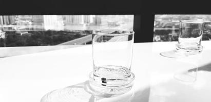 Closeup clear glass of water on white table and sunlight from side with window, cityscape view and transportation in city background in black and white style. Object and Art in monochrome tone. photo