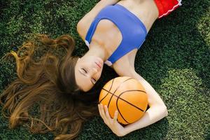 Attractive athletic young woman posing outdoors, lying on her back on the lawn and holding a basketball photo