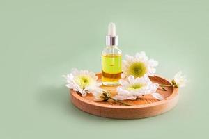 a glass bottle with a pipette and a white cap with a natural cosmetic, serum or oil stands on a wooden plate with flowers. green background. photo