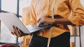 mode ontwerper l jong Aziatisch vrouw werken gebruik makend van laptop, tablet en glimlachen terwijl staand in werkplaats reageren Aan bedrijf video